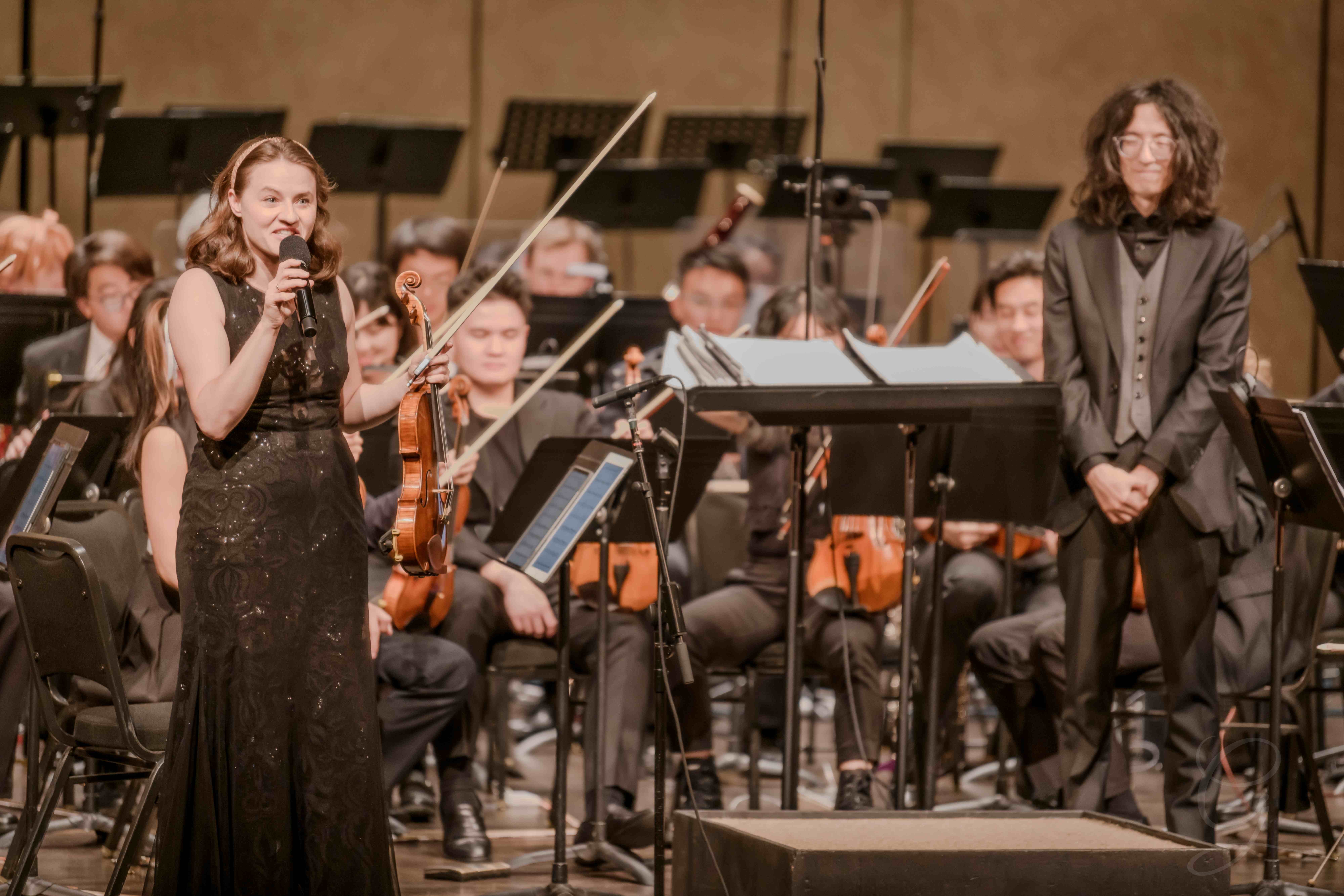 Vocalist speaking to the audience with her violin as the orchestra looks on